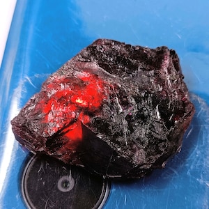 May include: A large, rough, dark purple and red gemstone with a deep red interior. The gemstone is sitting on a blue surface.