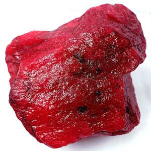 May include: A rough, uncut ruby gemstone. The ruby is a deep, saturated red colour with a rough, textured surface. Darker inclusions are visible throughout the stone. The gemstone is displayed against a white background.