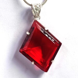 以下が含まれることがあります： 銀色の金属製の爪留め枠にセットされた、大きな凧型のファセットカットが施された赤い宝石のペンダント。ペンダントの上部には、銀色のチェーンに繋がる装飾的なフィリグリースタイルの金属製バチカンが付いています。