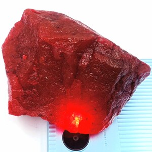 May include: A large, rough, red ruby gemstone. The ruby is a deep red color and has a natural, unpolished finish.