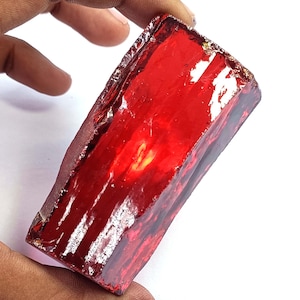 May include: A large, rectangular, translucent red gemstone with a polished, glossy surface and visible internal fractures. The stone displays deep crimson hues and sharp, angular edges, showcasing a raw yet refined crystalline structure.