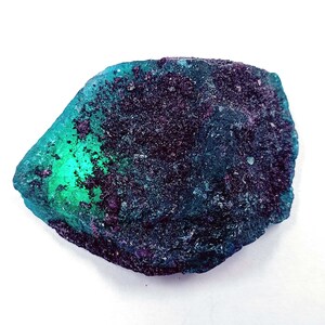 May include: A teal blue and black gemstone with a rough, textured surface. The gemstone has a green hue in the center and is irregularly shaped.