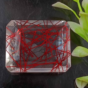 May include: A clear quartz crystal with red inclusions. The crystal is cut into a rectangular shape with beveled edges. The red inclusions are arranged in a random pattern throughout the crystal.