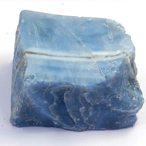 May include: A raw, uncut piece of blue opal gemstone. The stone displays a range of blue hues, from light sky blue to deeper shades, with visible natural textures and a subtle white band running across the center. The opal is a natural mineral.