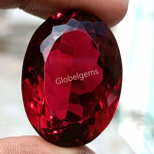 May include: A large, oval-shaped, red gemstone with a faceted cut. The gemstone is held in a hand and the word "Globelgems" is visible in the center of the stone.