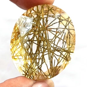 May include: Oval-shaped, clear quartz gemstone with gold rutile inclusions. The gemstone has a translucent appearance with a high concentration of gold rutile needles.