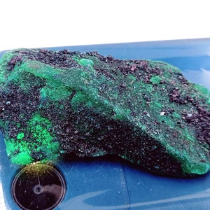 May include: A rough, green and black rock with a textured surface. The rock has a dark green hue with black specks throughout.