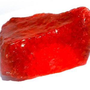 Puede incluir: Un espécimen mineral rojo, bruto y translúcido, con una forma irregular y dentada, y una textura superficial cristalina y brillante. La piedra muestra tonos variados de carmesí y escarlata vibrantes, con facetas naturales visibles.