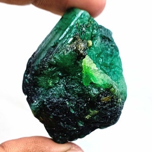 May include: A large, rough, green emerald gemstone with a dark green, almost black, exterior. The emerald has a natural, unpolished look.