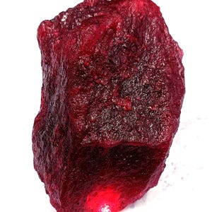 Op de afbeelding: Een ruwe, ongeslepen robijn edelsteen met een dieprode kleur. Het oppervlak heeft een textuur, ongelijkmatig uiterlijk, met gebieden van licht en schaduw. De edelsteen is een natuurlijk, ruw mineraal.