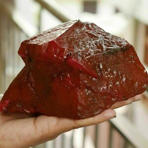 May include: A large, rough-cut ruby gemstone with a deep red color. The raw mineral is held in a hand, showcasing its irregular shape and natural texture. The gemstone appears to be uncut and unpolished.