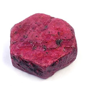 May include: A raw, hexagonal-shaped ruby crystal with a deep magenta-red colour. The surface of the gemstone displays a rough, textured appearance with visible dark inclusions and natural crystalline facets across its top and sides.