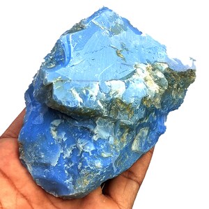 May include: A large, raw chunk of blue opal mineral with a jagged, uneven surface. The stone displays a vibrant, opaque sky-blue color with patches of white and earthy brown inclusions embedded within the crystalline, fractured rock structure.