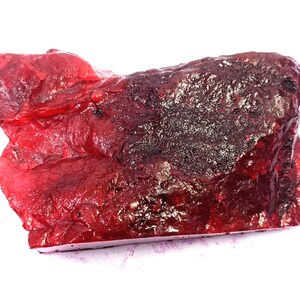 Könnte beinhalten: Ein rohes, unpoliertes Mineralexemplar mit einer leuchtend durchscheinenden roten Farbe. Der Stein weist eine unregelmäßige, gezackte Oberfläche mit tiefroten und dunkel kastanienbraunen Variationen sowie eine kristalline Textur auf.