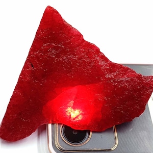 May include: A large, rough, red gemstone with a triangular shape. The gemstone is illuminated from below, causing it to glow.