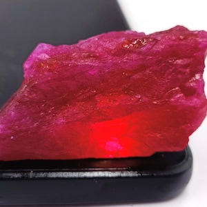 May include: A raw, unpolished ruby gemstone with a vibrant, deep pinkish-red hue. The stone features a jagged, crystalline texture with translucent areas that catch the light, displaying natural facets and irregular edges across its surface.