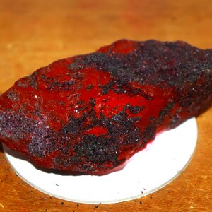 Peut inclure: Un spécimen minéral brut et allongé présentant une surface rouge vif et brillante contrastée par des zones de dépôts noirs cristallins et texturés. La pierre irrégulière et dentelée repose sur une base circulaire blanche.