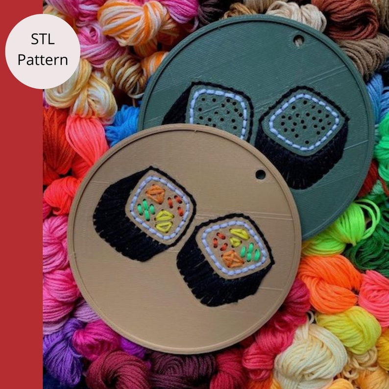 K&ouml;nnte beinhalten: Zwei braune Holzkreise mit einem Loch oben. Jeder Kreis hat ein gesticktes Design von zwei Sushi-Rollen. Die Sushi-Rollen sind schwarz, wei&szlig;, gr&uuml;n und orange. Die Kreise sind von buntem Garn umgeben.