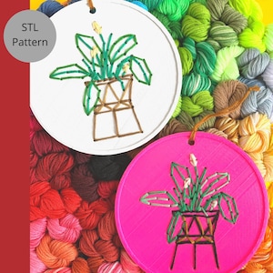 Puede incluir: Dos aros de bordado con un diseño de planta. Un aro es blanco con una planta verde y una maceta marrón. El otro aro es rosa con una planta verde y una maceta marrón.