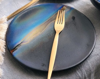 May include: A black ceramic dinner plate with a blue and gold iridescent glaze. A gold fork is placed on the plate.