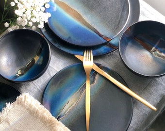May include: A set of four black and blue ceramic plates and bowls with a gold fork and knife. The plates and bowls have a unique iridescent glaze.
