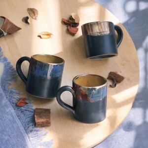 May include: Three ceramic mugs with dark blue exteriors and contrasting interior colors. The rims feature a blend of blue, brown, and gold tones. Each mug has a curved handle. The mugs are arranged on a wooden surface with decorative elements.