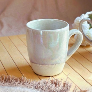 May include: A handmade ceramic mug with a pearlescent glaze. The mug has a rounded body and a sturdy handle. The bottom half of the mug is a matte cream color. The mug is sitting on a wooden surface.