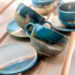 May include: A set of handmade ceramic teacups and saucers in a teal glaze with iridescent accents. The cups have a rounded shape with a handle, and the saucers are round. The set is arranged on a wooden surface.