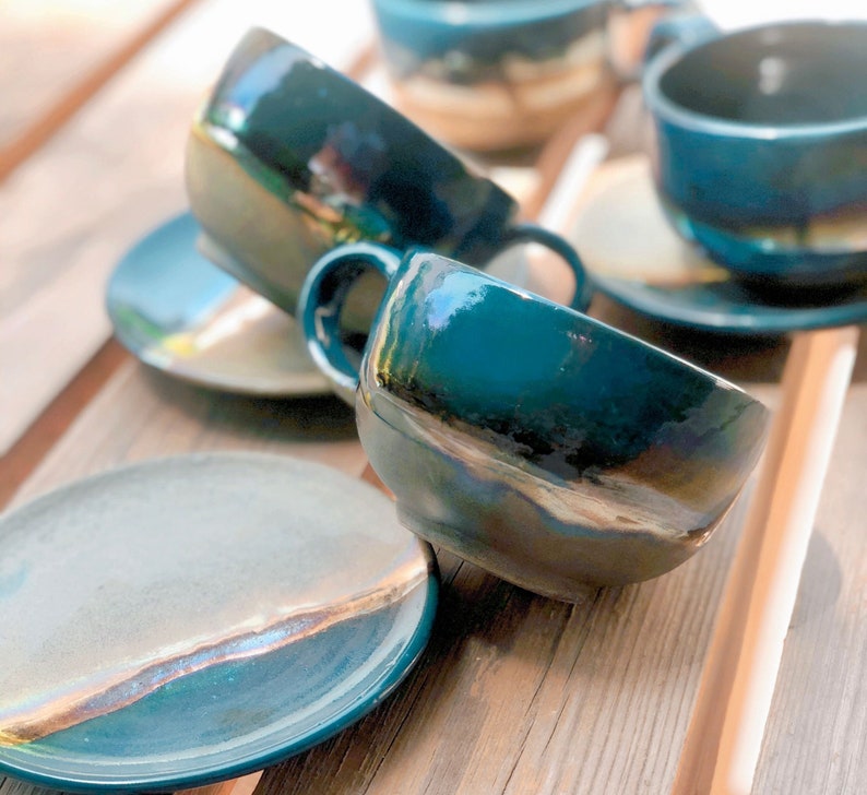 May include: A set of handmade ceramic teacups and saucers in a teal glaze with iridescent accents. The cups have a rounded shape with a handle, and the saucers are round. The set is arranged on a wooden surface.