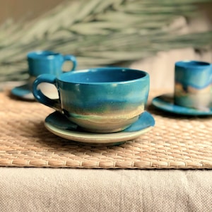 May include: Handmade ceramic teacups and saucers in shades of teal, blue, and green. The teacups have a rounded shape with a handle, and the saucers are circular. The glaze creates a layered effect, with a gradient of colors. The set is placed on a woven mat.