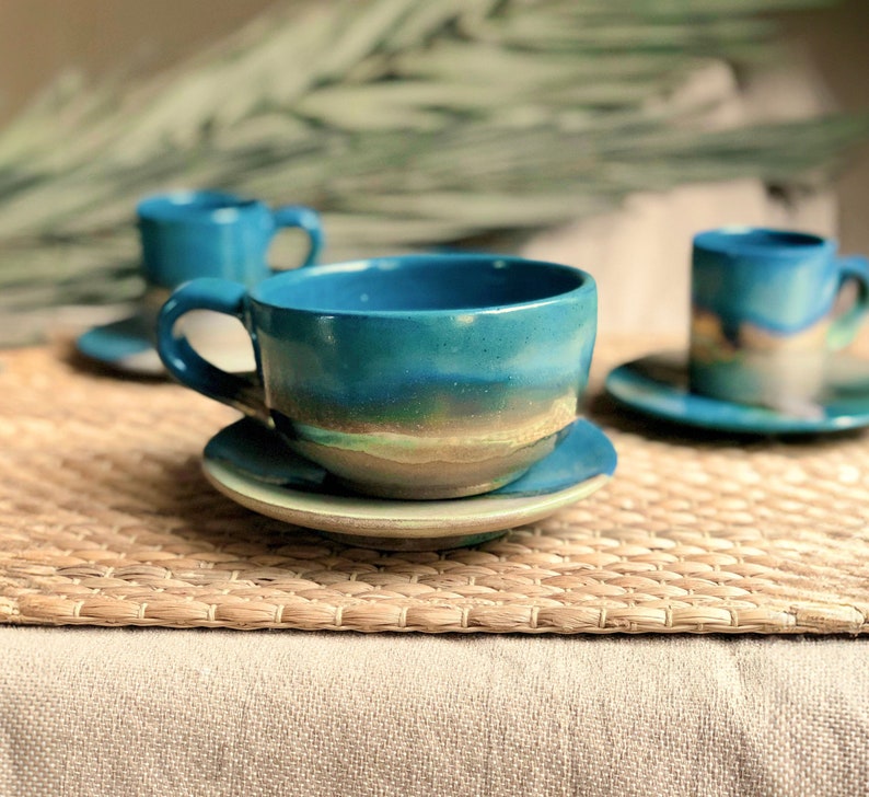 May include: Handmade ceramic teacups and saucers in shades of teal, blue, and green. The teacups have a rounded shape with a handle, and the saucers are circular. The glaze creates a layered effect, with a gradient of colors. The set is placed on a woven mat.