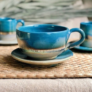 May include: Handmade ceramic teacups and saucers in a vibrant teal glaze. The cups have a rounded shape with a handle, and the saucers are a matching color. The set is displayed on a woven mat, creating a rustic and artistic aesthetic.