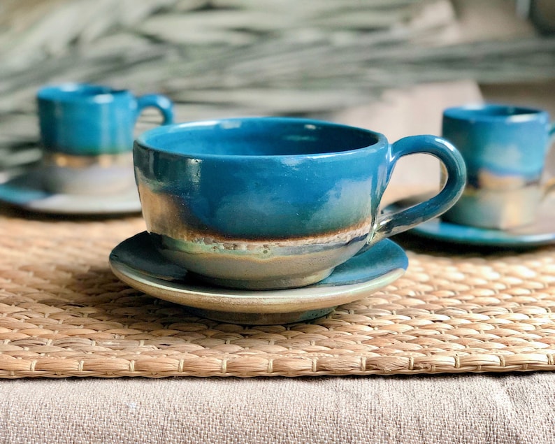 May include: Handmade ceramic teacups and saucers in a vibrant teal glaze. The cups have a rounded shape with a handle, and the saucers are a matching color. The set is displayed on a woven mat, creating a rustic and artistic aesthetic.