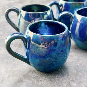 Può includere: Una collezione di tazze in ceramica fatte a mano con una smaltatura blu iridescente lucida. Ogni tazza ha un corpo arrotondato, un manico robusto e una variazione di colore unica. Le tazze sono disposte su una superficie grigia.
