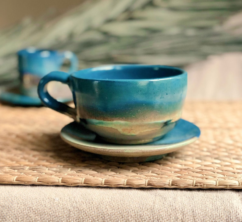May include: A handmade ceramic teacup and saucer set. The cup and saucer are a vibrant teal color with a gradient effect, transitioning to a lighter tan near the base. The cup has a rounded handle. A second cup is slightly visible in the background.