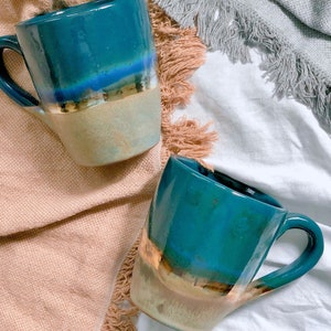 May include: Two ceramic mugs with a teal glaze and gold accents. The mugs have a unique, hand-painted design with a layered effect of blue, gold, and brown.