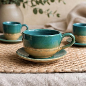 May include: A set of teal and tan ceramic teacups and saucers. The cups have a rounded shape with a handle and sit on matching saucers. The cups and saucers are placed on a woven mat, with a neutral background.