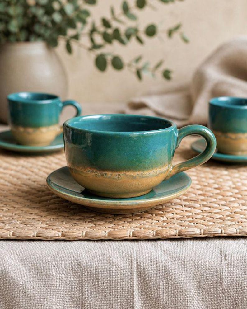 May include: A set of teal and tan ceramic teacups and saucers. The cups have a rounded shape with a handle and sit on matching saucers. The cups and saucers are placed on a woven mat, with a neutral background.