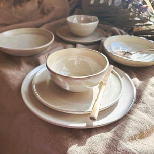 May include: A selection of beige ceramic tableware, including plates, bowls, and a small cup, arranged on a textured, neutral-toned surface. A golden knife rests on one of the plates. The set is bathed in natural light, creating a warm, inviting aesthetic.