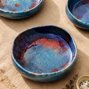 Może przedstawiać: Zestaw trzech ręcznie robionych ceramicznych misek z unikalnym, teksturowanym szkliwem. Miski charakteryzują się mieszanką odcieni niebieskiego, czerwonego i fioletowego, tworząc żywy, artystyczny wygląd. Miski są okrągłe z lekko nieregularnymi krawędziami.