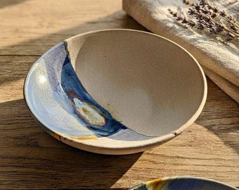 May include: A shallow ceramic bowl with a textured, off-white interior and a striking blue, brown, and gold glazed accent sweeping across one side. The bowl features a curved rim and a speckled finish, showcasing a contrast between the matte clay.
