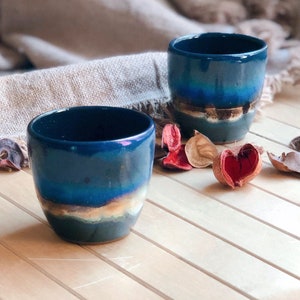 Op de afbeelding: Twee blauwe keramische mokken met een lichtblauwe en gouden glazuur. De mokken staan op een houten oppervlak met gedroogde bloemblaadjes.