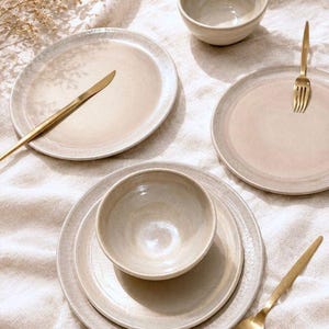 May include: A neutral-toned ceramic dinnerware set with plates, bowls, and gold-colored utensils. The plates have a slightly textured rim. A knife, fork, and spoon are arranged on the plates. The set is displayed on a light-colored, textured surface.
