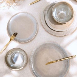 Könnte beinhalten: Eine Sammlung von Keramikgeschirr in neutralen Farbtönen, darunter Teller, Schalen und goldfarbenes Besteck. Das Set umfasst verschiedene Tellergrößen, eine Schüssel, eine Gabel, einen Löffel und ein Messer. Das Geschirr ist auf einer hellen Oberfläche angeordnet.