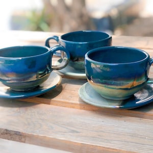 May include: Three ceramic teacups and saucers in a deep teal glaze with iridescent highlights. The cups have rounded bodies, large handles, and sit on matching saucers. The set is arranged on a wooden surface, suggesting a rustic or handmade aesthetic.