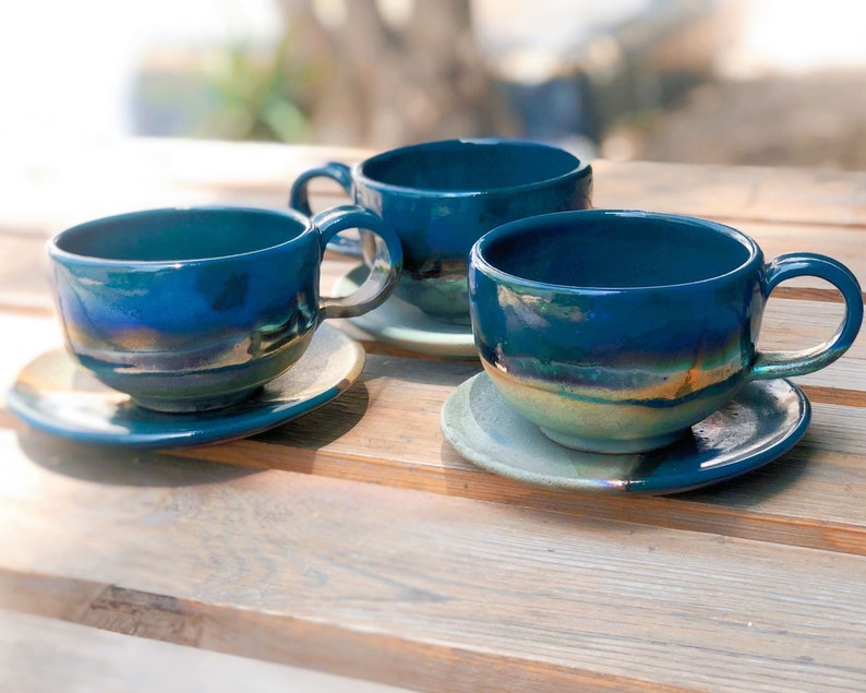May include: Three ceramic teacups and saucers in a deep teal glaze with iridescent highlights. The cups have rounded bodies, large handles, and sit on matching saucers. The set is arranged on a wooden surface, suggesting a rustic or handmade aesthetic.