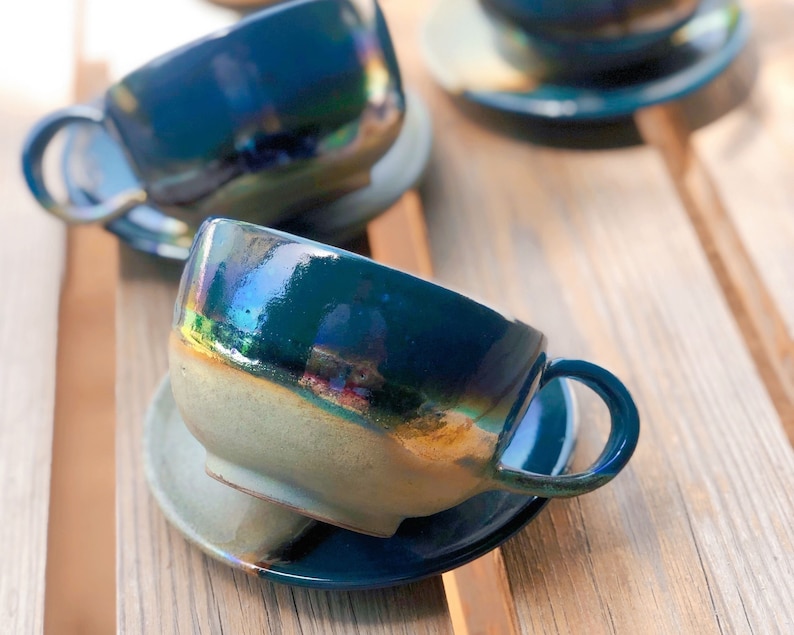 May include: A set of ceramic teacups and saucers. The cups are a deep teal color with iridescent highlights, and the saucers are a lighter, muted green. The cups have a rounded shape with a handle. The set is placed on a wooden surface.