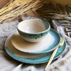 May include: A ceramic dinnerware set featuring a bowl and two plates with a speckled, off-white center and a teal blue rim. A gold fork rests on the side. The set is arranged on a gray cloth, with dried plants in the background.