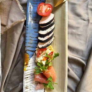 Può includere: Un piatto da portata rettangolare in ceramica con un design blu e beige, contenente una varietà di alimenti. Pomodori a fette, cibi a strati bianchi e neri e altri ingredienti sono disposti sul piatto.