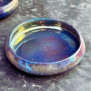 May include: A close-up of a small, round ceramic bowl with a metallic, iridescent finish. The bowl's colors shift from deep blues and purples to hints of gold and green. The bowl has a wide, shallow design.